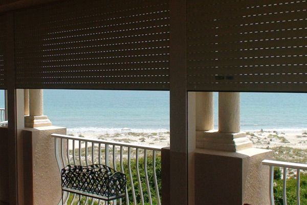 Beige window shutters partially obscuring a beach view. White columns and balcony railing frame the scene.