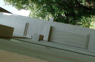 White building exterior with two rectangular louvered vents, angled view. Green foliage in the background.