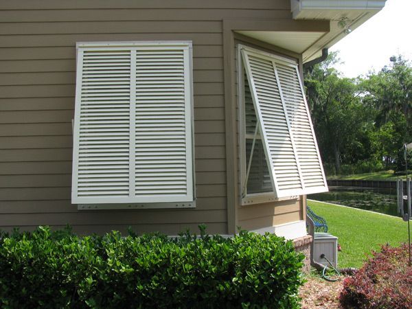 White louvered shutters on a brown house. One is closed, the other open. Green bushes in the foreground.
