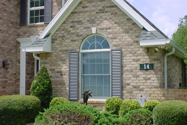 Brick house with arched window, shutters, and well-manicured landscaping; a dark cat is in the window.