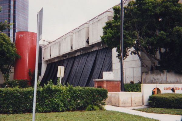 Building with red cylinder and black draping, trees, and sign in front of green shrubs.