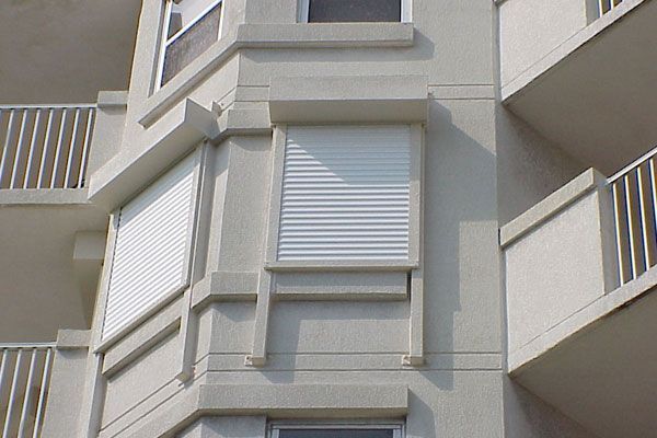 Corner of a building with white shuttered windows. Light gray exterior, balconies with white railings.