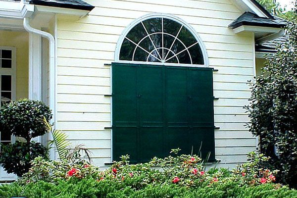 Green hurricane shutters cover a large, arched window on a yellow house.