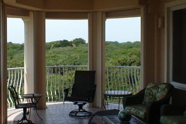 Interior of a room with a view of a lush green landscape through large windows and a balcony. Furnished with chairs and tables.