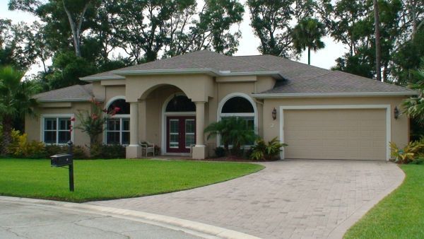 Beige stucco house with arched entryway, driveway, and garage door; green lawn and trees.