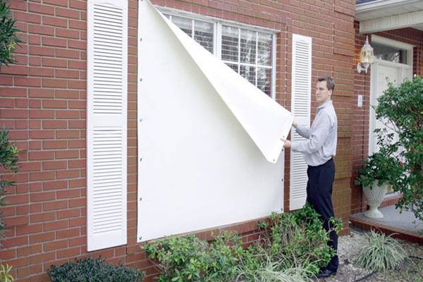 Man installing a white window cover on a brick house with white shutters.