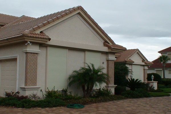 Beige house with light green shutters, brown tiled roof, and a cloudy sky.