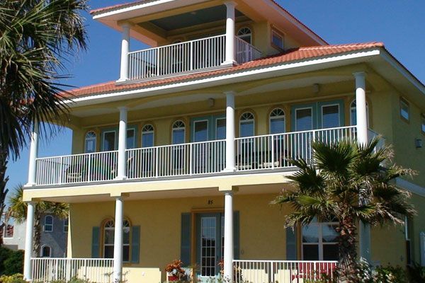 Yellow three-story beach house with white railings, columns, and a red tile roof. Palm trees in front.