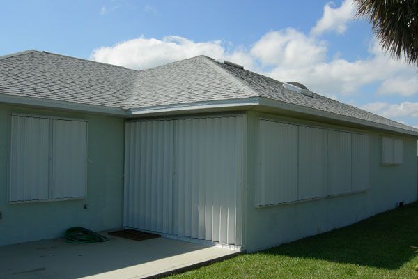 Exterior of a light green house with white hurricane shutters closed on several windows.