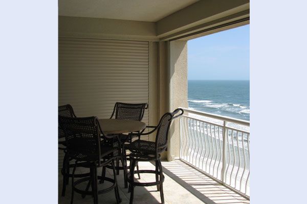Balcony with ocean view, table, chairs, and closed shutters. Sunny day, white railing.
