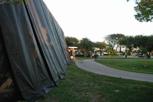 Dark tarp partially obscures a park scene with a paved path, trees, and people walking.