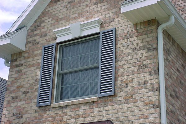 Brick house with gray shutters on a window, topped by white decorative trim.