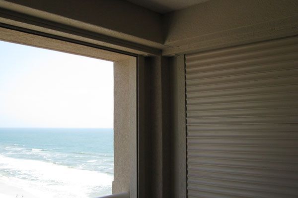 Ocean view from a building window, partial roller shutter, light blue sky and water.