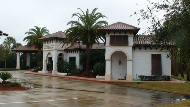 White stucco building with red tile roof, arched entryway, palm trees, and overcast sky.