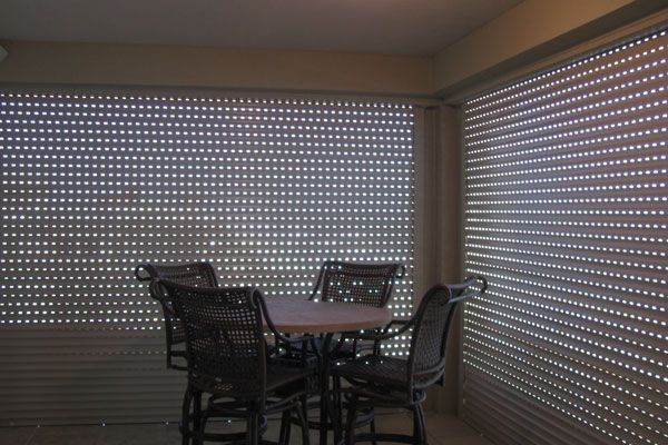 Outdoor seating area with round table, chairs, and light-filtering blinds.