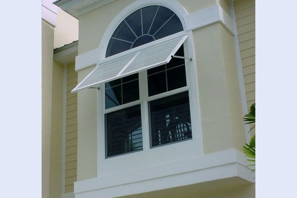 A white louvered awning over a rectangular window with an arched window above, on a yellow building.