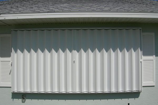 Accordion-style white hurricane shutters cover a window on a light green house.