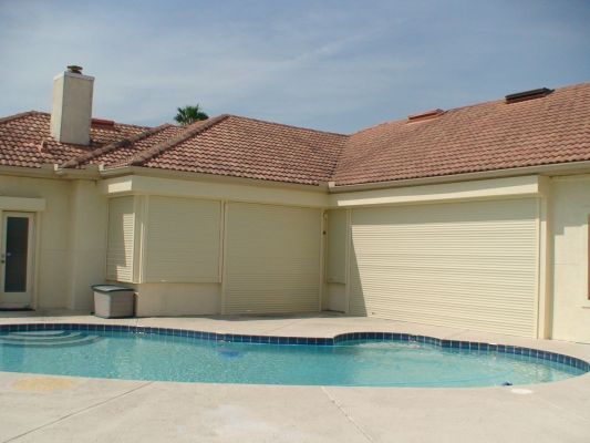 Swimming pool in front of a tan house with a red-tiled roof, with closed cream-colored shutters.