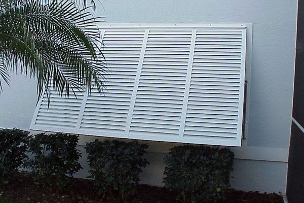 White, horizontal louvered window shutter, angled open, mounted on exterior wall above bushes.
