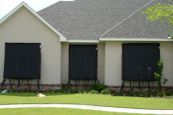 House with three windows covered by black hurricane shutters supported by metal stands, set in green lawn.