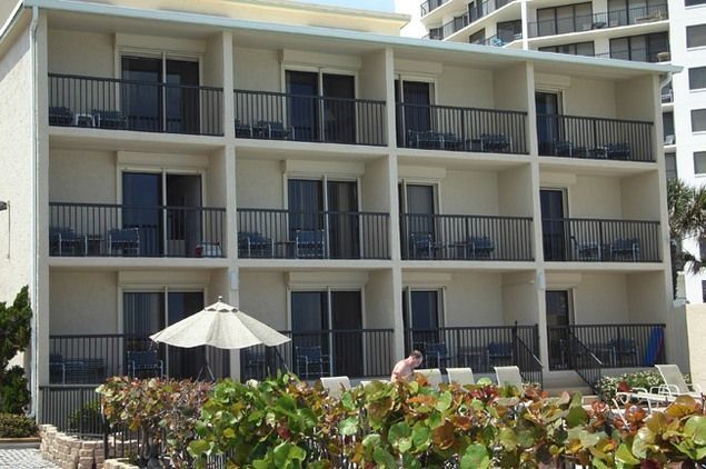 Two-story beachfront hotel with balconies, dark railings, and a neutral color scheme.