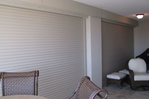 Beige roll-down shutters cover patio openings, with wicker furniture in the foreground.