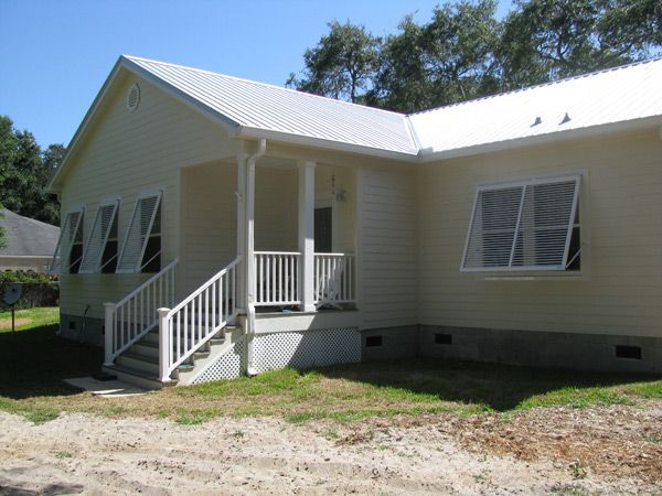 Yellow house with white shutters, porch, stairs, and metal roof against a blue sky.