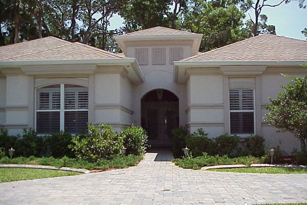 Beige stucco house with brick walkway, arched doorway, and shuttered windows. Lush greenery.