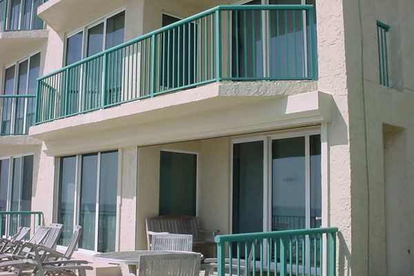 Exterior of a beige building with turquoise railings and sliding glass doors, overlooking a beach.
