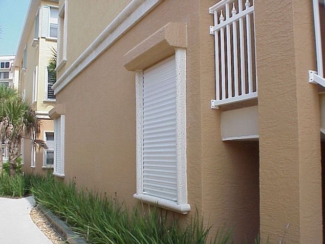Tan building exterior with white hurricane shutters on windows, green plants along the base.