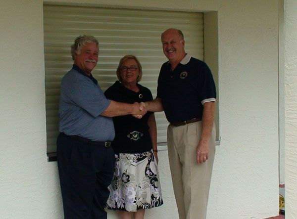Three people shaking hands in front of a window. Man in blue shirt, woman, man in khaki pants.