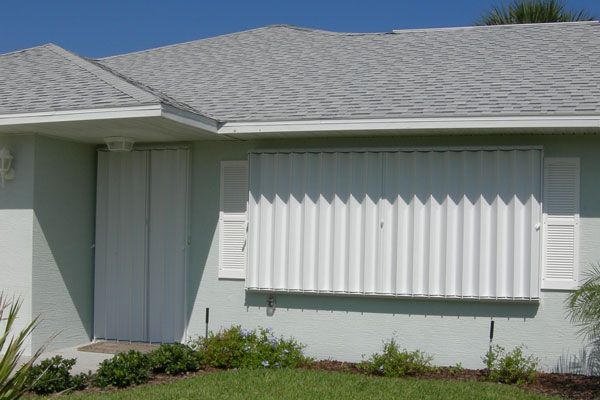 House with white hurricane shutters closed on door and window, green facade, gray roof.