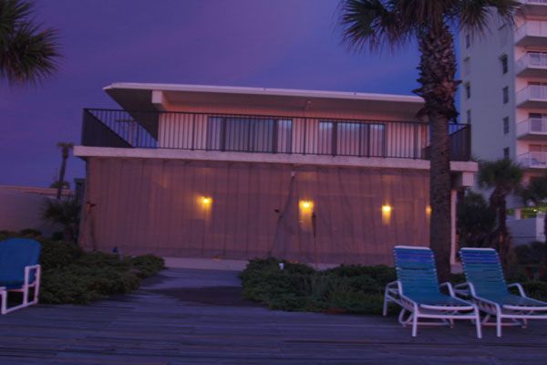 A modern beachside building with a second-floor balcony, surrounded by palm trees and lounge chairs at dusk.