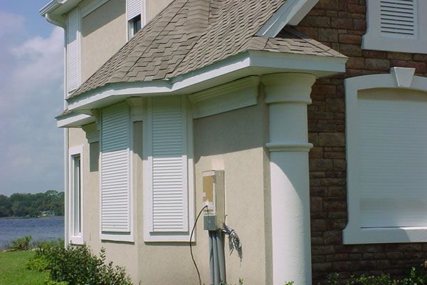 House exterior with closed white hurricane shutters, stucco and brick, near water.
