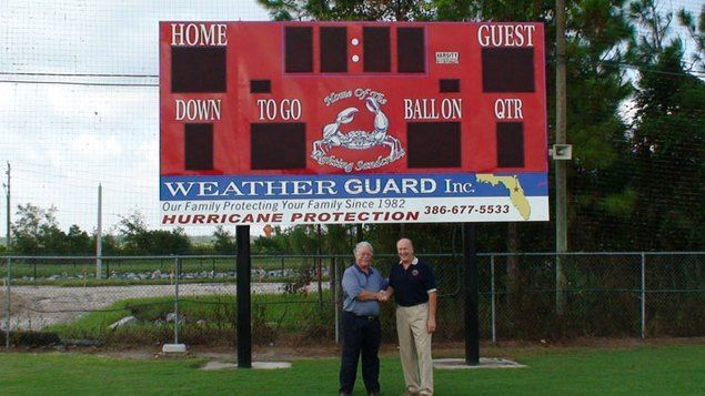 Two men stand in front of a red football scoreboard, below a Weather Guard sign.