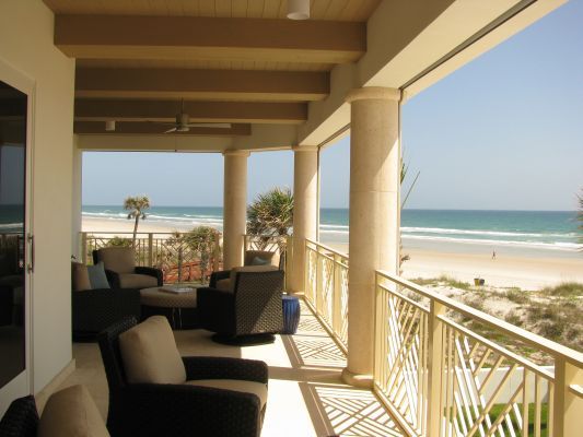 Balcony overlooking beach with ocean. Columns, railing, and seating on patio; trees and people on the sand.