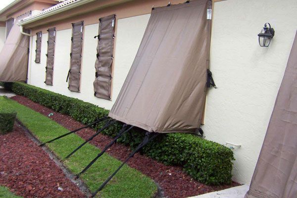 Brown hurricane shutters covering windows on a light beige building. Green hedge and red mulch in front.