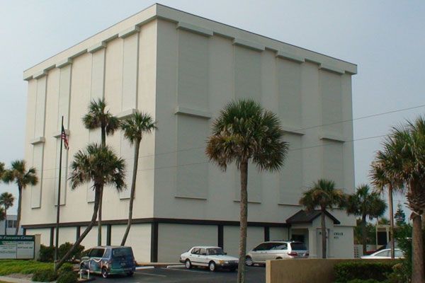 White, boxy building with palm trees in front; parking lot with cars.