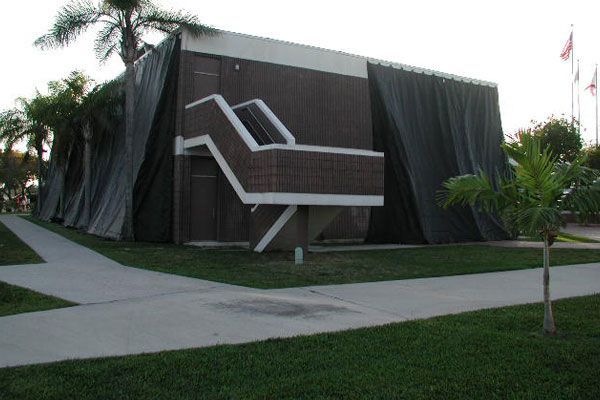 Building with exterior stairs and dark draping. Sidewalk, palm trees, and flags visible.