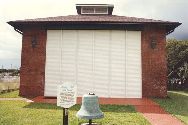 Brick building with white shutters, a bell in front, and a sign.