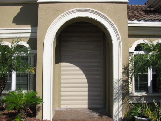 Tan roll-down shutter in an arched doorway of a tan stucco building with white trim.