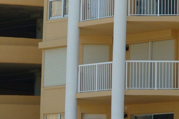 Exterior view of a beige building with white balconies and pillars; some windows have closed white shutters.