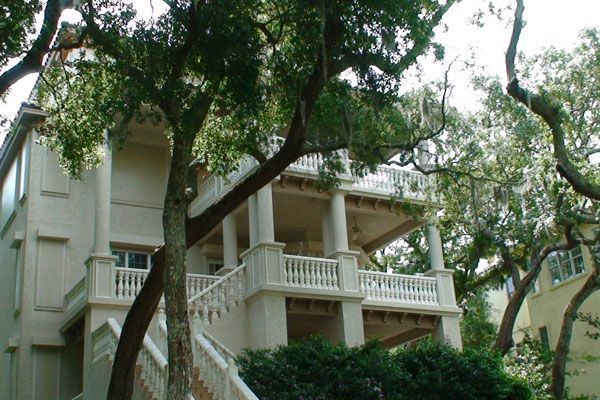 Beige two-story house with a white balcony and porch, partly obscured by tree branches.