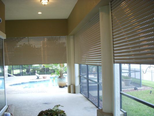 Beige roll-down shutters cover large openings of a patio overlooking a pool and yard.