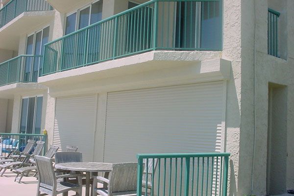 Exterior of a beachfront building with balcony and closed white roll-down shutters. Turquoise railing surrounds balcony.