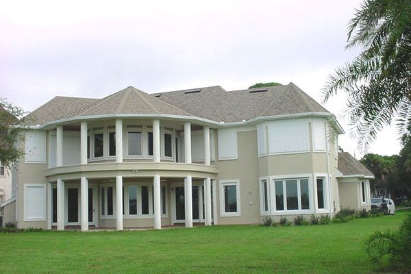 Two-story beige house with white columns, lawn, and storm shutters.