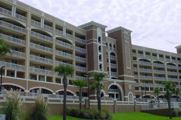 Multi-story beige building with balconies, palm trees, and green grass against a cloudy sky.