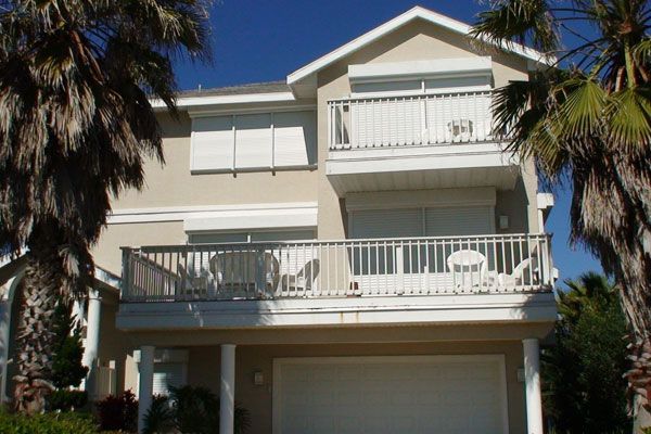 Two-story beige house with balconies, roll-down shutters, and palm trees. Garage door below.
