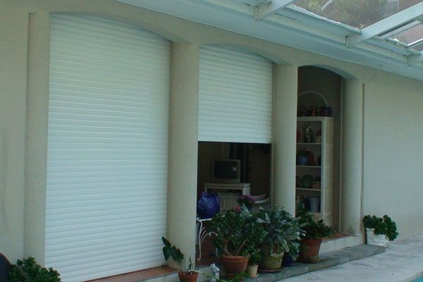 White rolling shutters on a building exterior, partially covering a window. Lush green plants are visible.