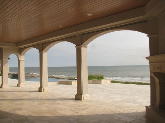 Arched patio overlooking ocean; beige columns, stone floor, wooden ceiling, overcast sky.